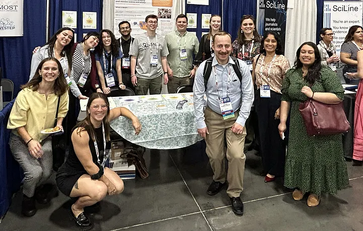 Several SNAP members gather around the expo booth table on opening night. All members are facing the camera and smiling as we celebrate SNAP’s presence at AAAS and get to meet each other after a year of virtual contact and meetings.