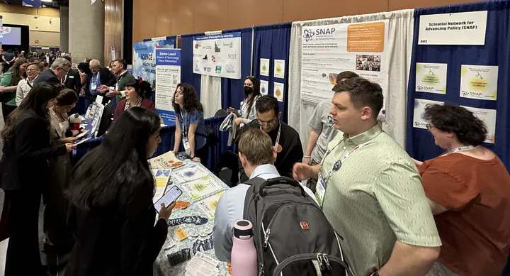Several people surround the SNAP expo booth. SNAP members interact with other AAAS conference attendees and answer questions about our mission and how other early career researchers can join.